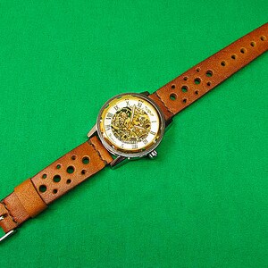 May include: A brown leather watch band with a silver and gold watch face. The watch face has Roman numerals and a skeleton design.