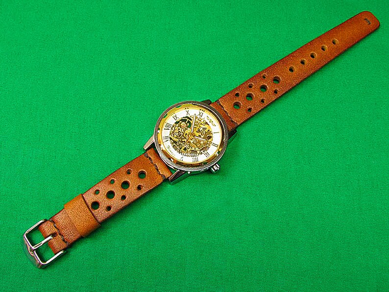 May include: A brown leather watch band with a silver and gold watch face. The watch face has Roman numerals and a skeleton design.