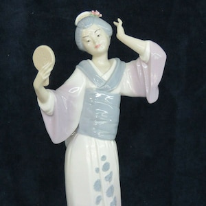 May include: A porcelain figurine of a woman in a traditional Japanese kimono. She is holding a hand mirror and has a flower in her hair. The kimono is white with blue and pink floral designs.