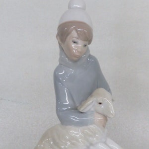 May include: A porcelain figurine of a child wearing a white hat and blue sweater holding a white lamb.