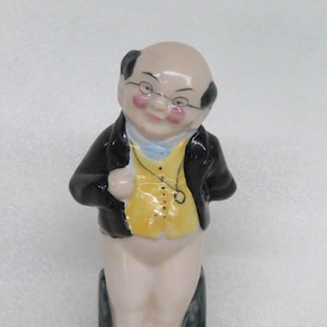 May include: A ceramic figurine of a man wearing a black jacket, yellow vest, and white pants. He has a bald head with black hair on the sides and is wearing glasses. He is smiling and has rosy cheeks.