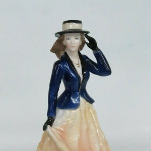 May include: A porcelain figurine of a woman wearing a blue jacket, a white blouse, a peach-colored skirt, and a black hat. She is holding her hat with her right hand.