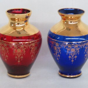 May include: Two decorative glass vases with gold accents. The vase on the left is red with gold floral designs. The vase on the right is blue with gold floral designs.