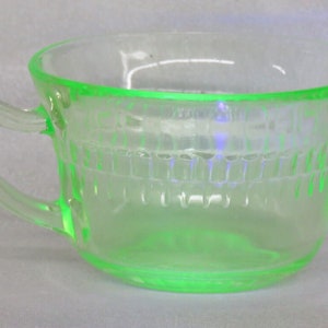 May include: A green glass cup with a handle. The cup has a textured design and is made of uranium glass.
