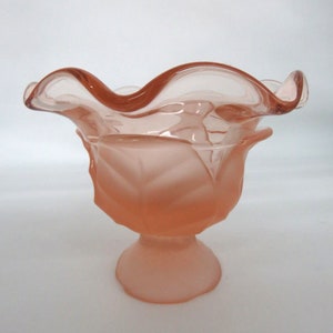 May include: A pink glass bowl with a ruffled edge and a frosted finish. The bowl is on a pedestal base.