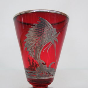 May include: A red glass goblet with a silver-plated rim and base. The goblet features a silver-plated design of a fish with a long, flowing tail.