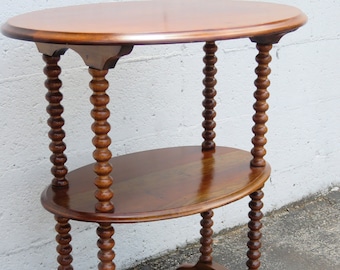 May include: A wooden oval-shaped table with two shelves. The table legs are made of turned wood with a beaded design. The table is brown and has a glossy finish.