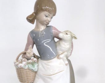 May include: A porcelain figurine of a young person holding a lamb and a basket of flowers. The figure wears a gray vest, pink sleeves, and a white apron. The base is a grassy green.