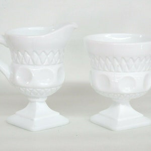 May include: A white milk glass creamer and sugar bowl set with a pedestal base and a diamond pattern. The creamer has a handle and a spout.