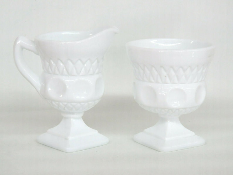 May include: A white milk glass creamer and sugar bowl set with a pedestal base and a diamond pattern. The creamer has a handle and a spout.