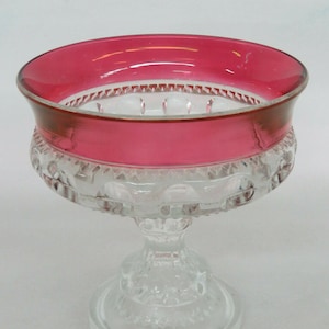May include: A clear glass pedestal bowl with a red rim. The bowl has a decorative pattern and a pedestal base.