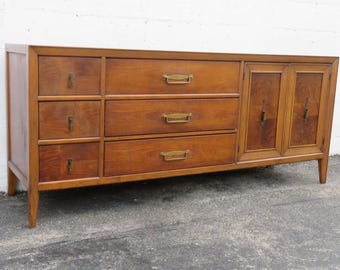 May include: A mid-century modern wooden dresser with a warm brown finish. It features six drawers with brass handles on the left and two cabinet doors on the right. The dresser is supported by tapered legs.
