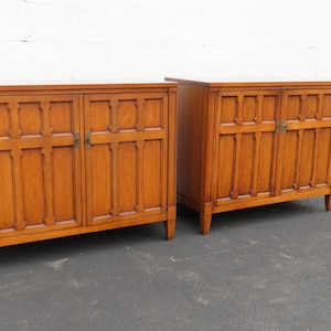 May include: Two matching wooden cabinets with two doors each. The cabinets are a warm brown color with vertical and horizontal carved details on the doors. Each cabinet has brass-colored hardware and sits on short, angled legs.