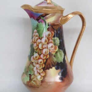 May include: A vintage porcelain pitcher with a gold trim and a painted design of grapes and leaves. The pitcher has a lid with a gold knob.
