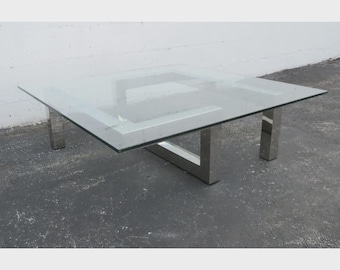 May include: A square coffee table featuring a clear glass top and a reflective chrome metal base. The base consists of four vertical legs connected by a horizontal frame structure that supports the glass surface from underneath.