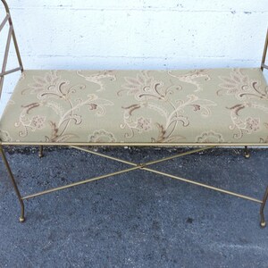 May include: A gold metal bench with a floral patterned cushion. The bench has a simple, elegant design with curved arms and a criss-cross base.