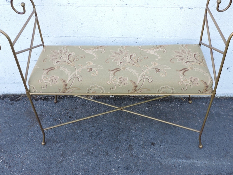 May include: A gold metal bench with a floral patterned cushion. The bench has a simple, elegant design with curved arms and a criss-cross base.