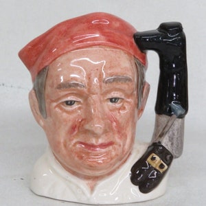 May include: A ceramic character mug depicting a man wearing a red hat and holding a brown boot in his hand. The mug is hand-painted and has a detailed design.