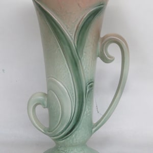 May include: A tall, ceramic vase with a flared top and two curved handles. The vase features a two-tone color scheme, with a pinkish-tan upper section and a light green lower section. Green leaf-like designs adorn the vase's exterior.
