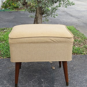 May include: A beige upholstered ottoman with wooden legs. The ottoman has a tufted top and a rectangular shape.