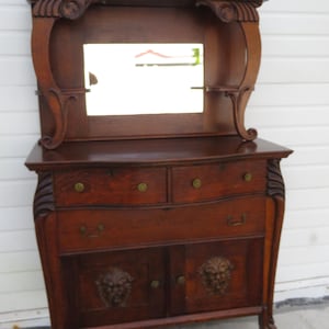 May include: A dark brown antique dresser with a mirror, drawers, and cabinet doors. The dresser has ornate carvings, including lion head details on the cabinet doors. The mirror is rectangular and set within a decorative frame.