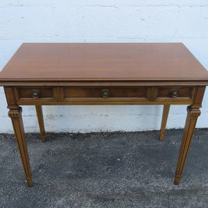 May include: A vintage wooden desk with three drawers and brass hardware. The desk has a rectangular top and four slender, tapered legs. The wood has a warm, brown tone, and the desk appears to be in good condition.