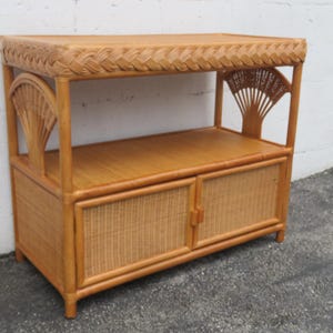 May include: A light brown rattan console table featuring a flat top with a braided edge, an open middle shelf, and a lower cabinet with two doors. The sides of the upper section are decorated with fan-shaped rattan accents and vertical supports.