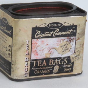May include: A rectangular metal Bigelow Constant Comment tea tin with a black lid and base. The cream-colored label features black text, a graphic of a teapot pouring into a cup, and the words Tea Bags, Flavored with rind of Oranges, and Sweet Spice.