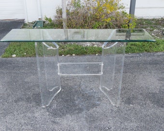 May include: A clear acrylic console table with a glass top and a lower shelf. The table has a modern design with curved legs.
