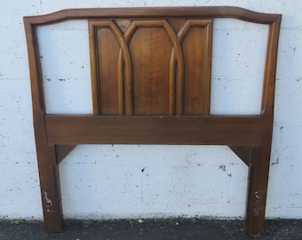 May include: A wooden headboard with a dark brown finish. The headboard features a rectangular design with an arched top and a central panel with three vertical, arched details. The headboard is supported by two legs.