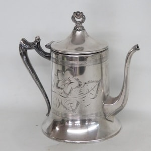 May include: A silver-toned metal teapot featuring a cylindrical body with a flared base, a curved handle, and a slender spout. The exterior surface is decorated with an engraved floral motif and horizontal line accents, topped with a lid featuring a small decorative finial.
