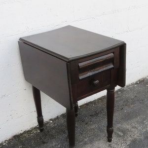 May include: A dark brown wooden drop leaf table with a single drawer and turned legs. The table is closed and the leaf is down.
