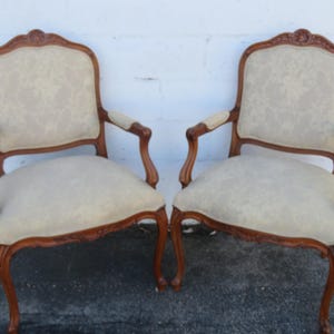 May include: Two antique armchairs with ornate wooden frames and light-colored patterned upholstery. The chairs have curved legs and armrests, showcasing a classic design. The wood has a warm, reddish-brown tone, and the fabric appears to be a neutral beige.