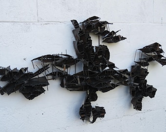 May include: Abstract metal wall sculpture made of many small pieces of metal welded together. The sculpture is black and has a jagged, organic shape.