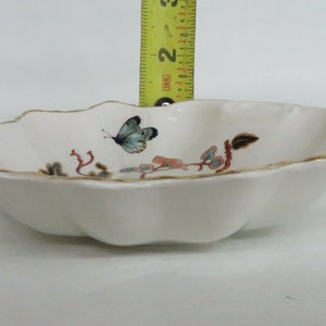 May include: A white porcelain bowl with a scalloped edge and a gold trim. The bowl features a hand-painted design of a butterfly and floral accents.