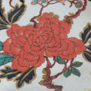 May include: A close-up of a floral design on a white ceramic plate. The design features a large, red flower with a swirling pattern, surrounded by green and blue leaves and branches.