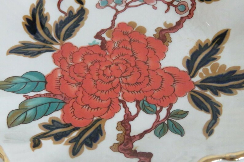 May include: A close-up of a floral design on a white ceramic plate. The design features a large, red flower with a swirling pattern, surrounded by green and blue leaves and branches.