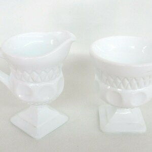 May include: White milk glass creamer and sugar bowl set with a raised, textured design. The creamer has a handle and a spout. The sugar bowl has a round top and a square base.
