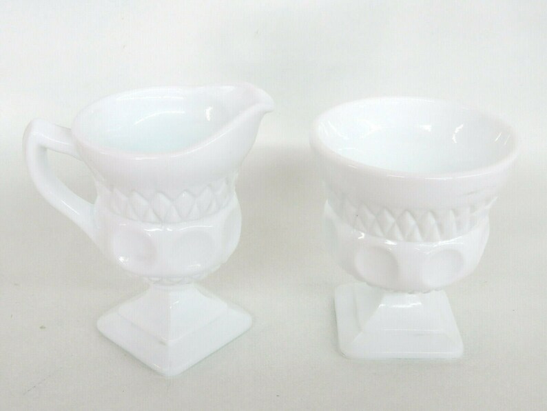 May include: White milk glass creamer and sugar bowl set with a raised, textured design. The creamer has a handle and a spout. The sugar bowl has a round top and a square base.