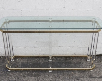 May include: A glass-top console table with a brass frame. The table has a rectangular top and four legs. The table is made of clear glass and brass.