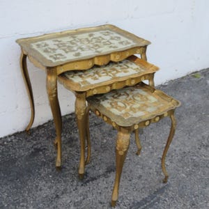 May include: A set of three gold-tone nesting tables with intricate floral designs on the tops. The tables have a distressed finish and are made of metal.