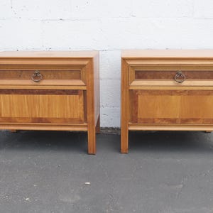 May include: A pair of light brown wooden nightstands, each featuring a single drawer with a circular brass pull. The drawer fronts and lower panels display a contrasting wood grain pattern with darker, mottled borders and lighter centers.