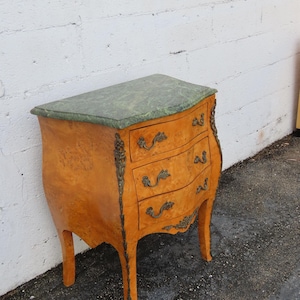 May include: A small, antique chest of drawers with a curved front and three drawers. The chest has a light brown, wood-grain finish, ornate brass hardware, and a green marble top. The legs are curved and the overall style is French Provincial.