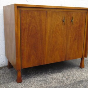May include: A mid-century modern wooden cabinet with two doors and small, dark-colored handles. The cabinet is a warm brown color and has a rectangular shape with four short legs. The cabinet is likely used for storage.