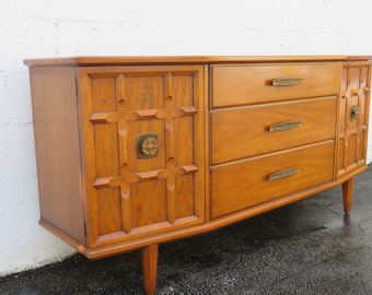 May include: A mid-century modern wooden dresser with three drawers and two cabinet doors. The cabinet doors have a carved geometric pattern and brass hardware. The dresser has a light wood finish and sits on four tapered legs.