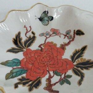 May include: A white porcelain dish with a scalloped edge and a gold trim. The dish features a floral design with a large red flower, green leaves, and a black butterfly.