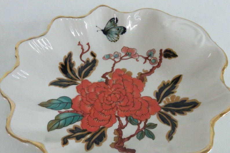 May include: A white porcelain dish with a scalloped edge and a gold trim. The dish features a floral design with a large red flower, green leaves, and a black butterfly.