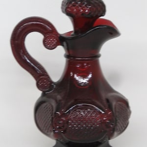 May include: A ruby red glass pitcher with a handle and a textured stopper. The pitcher has a decorative pattern and a spout for pouring. The handle has a heart-shaped design. The pitcher is likely for serving beverages.