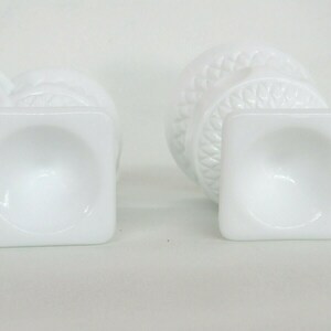 May include: Two white milk glass vases with a square base and a decorative design. The vases are empty and have a smooth, glossy finish.