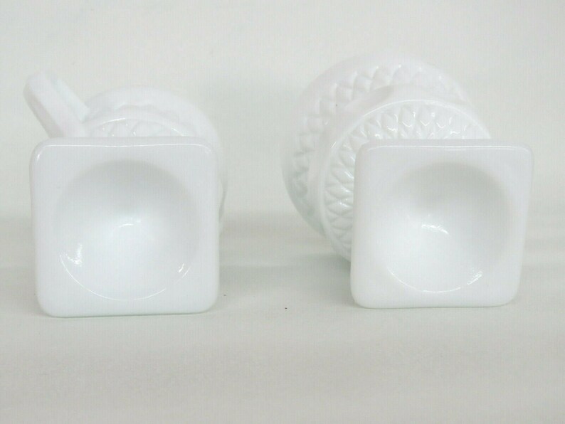May include: Two white milk glass vases with a square base and a decorative design. The vases are empty and have a smooth, glossy finish.